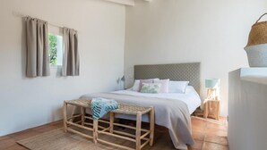 Ein Schlafzimmer mit Bett, Nachttisch, Lampe, Fenster mit Vorhängen und einem gewebten Hocker.