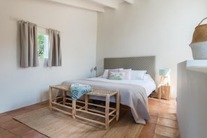 A bedroom with a bed, a bedside table, a lamp, a window with curtains, and a woven stool. - Can Busquera (Sóller)