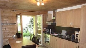 A wooden interior with a dining table, chairs, a kitchen with wooden cabinets, and a stone wall visible through the door.
