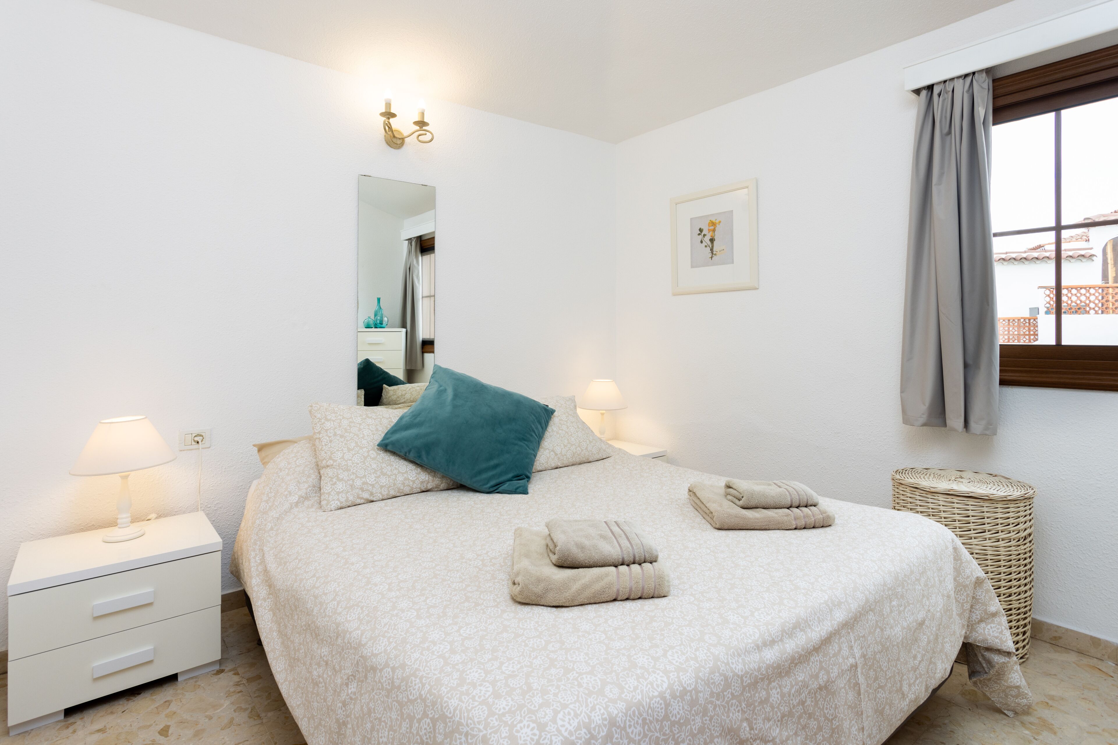 Un dormitorio ordenado con una cama, dos mesitas de noche, un espejo, un cuadro en la pared y una ventana con cortinas.