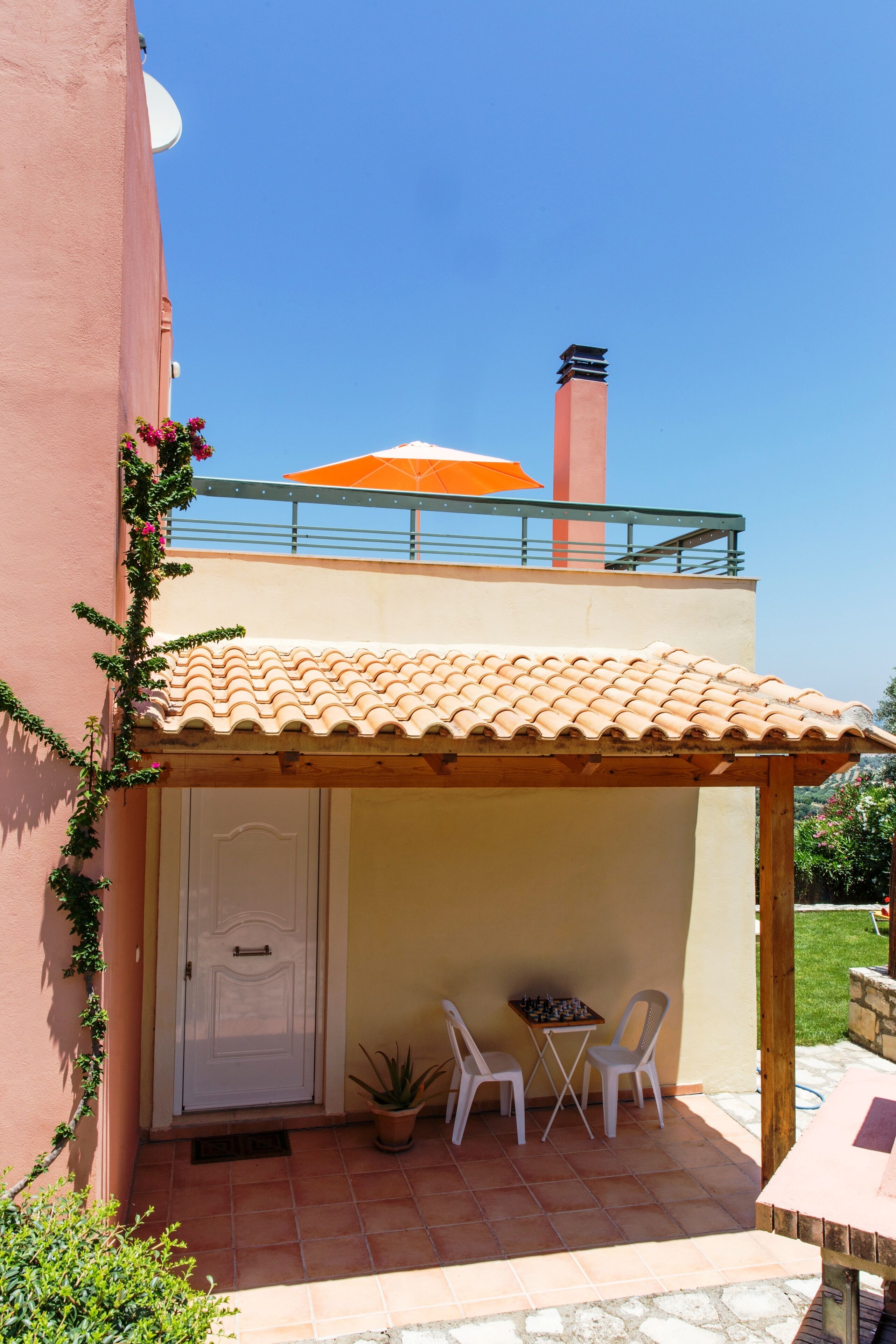 A two-story building with a tiled roof, a balcony with an orange umbrella, a door, and two chairs under a wooden pergola.