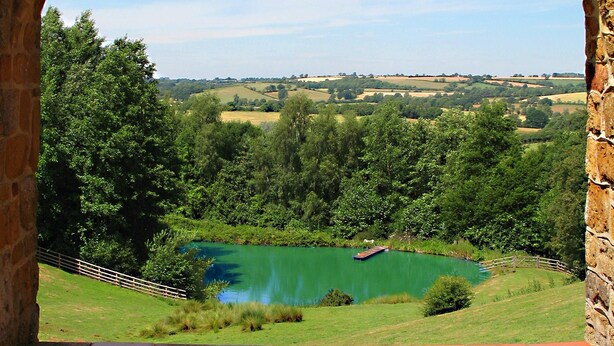 Un arc en pierre surplombe un paysage verdoyant, avec un lac et des champs qui s’étendent à l’horizon.