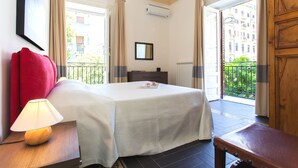 Een hotelkamer met een bed, nachtkastje met lamp, dressoir en balkon met uitzicht op gebouwen.