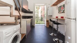 Een compacte keuken met een wasmachine, een klein eethoekje met twee krukken, en uitzicht op een balkon.