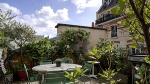 A patio with a round table and chairs, surrounded by greenery and a brick building. - CHARMING, AIR-CONDITIONED HOUSE WITH ROOFTOP, PARIS, CANAL ST. MARTIN (Paris)