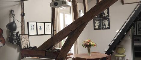 Une pièce avec un plafond haut, des poutres apparentes, un canapé, une table, une chaise, un piano et un escalier.