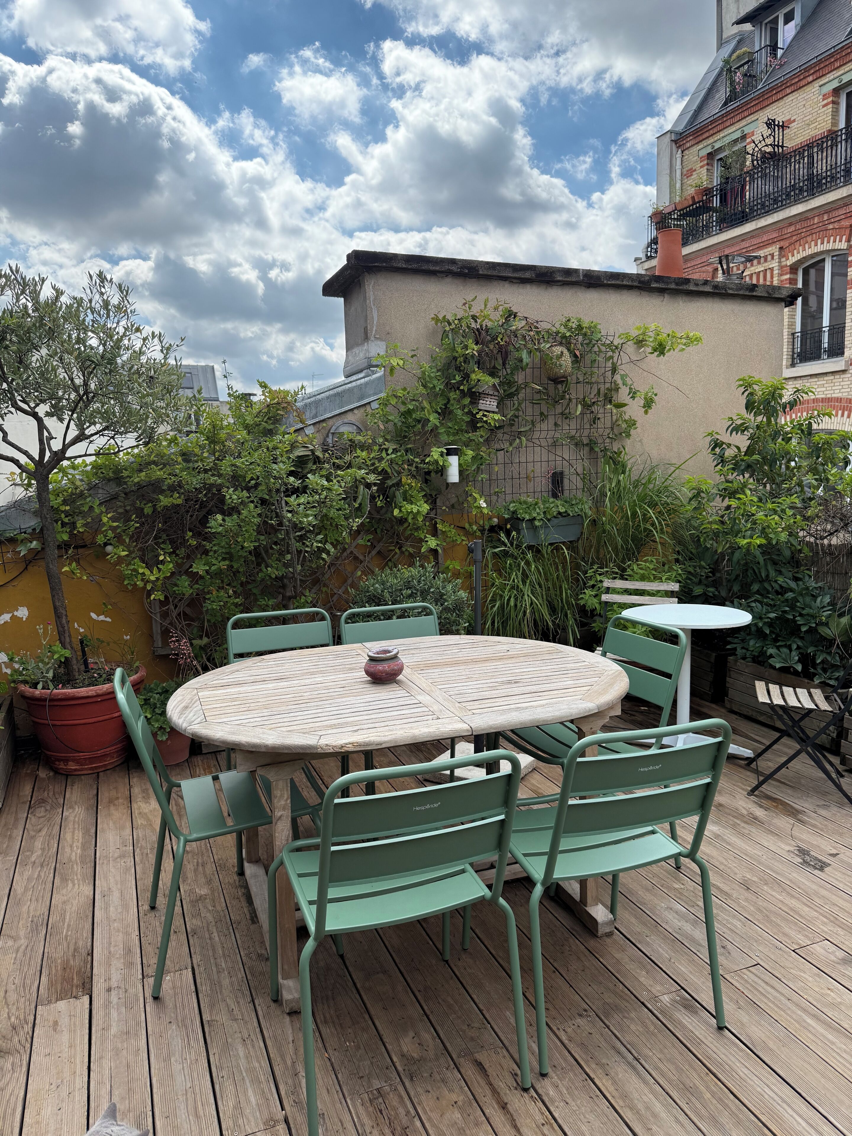 A wooden deck with a round table and green chairs, surrounded by potted plants and a building with a brick facade.