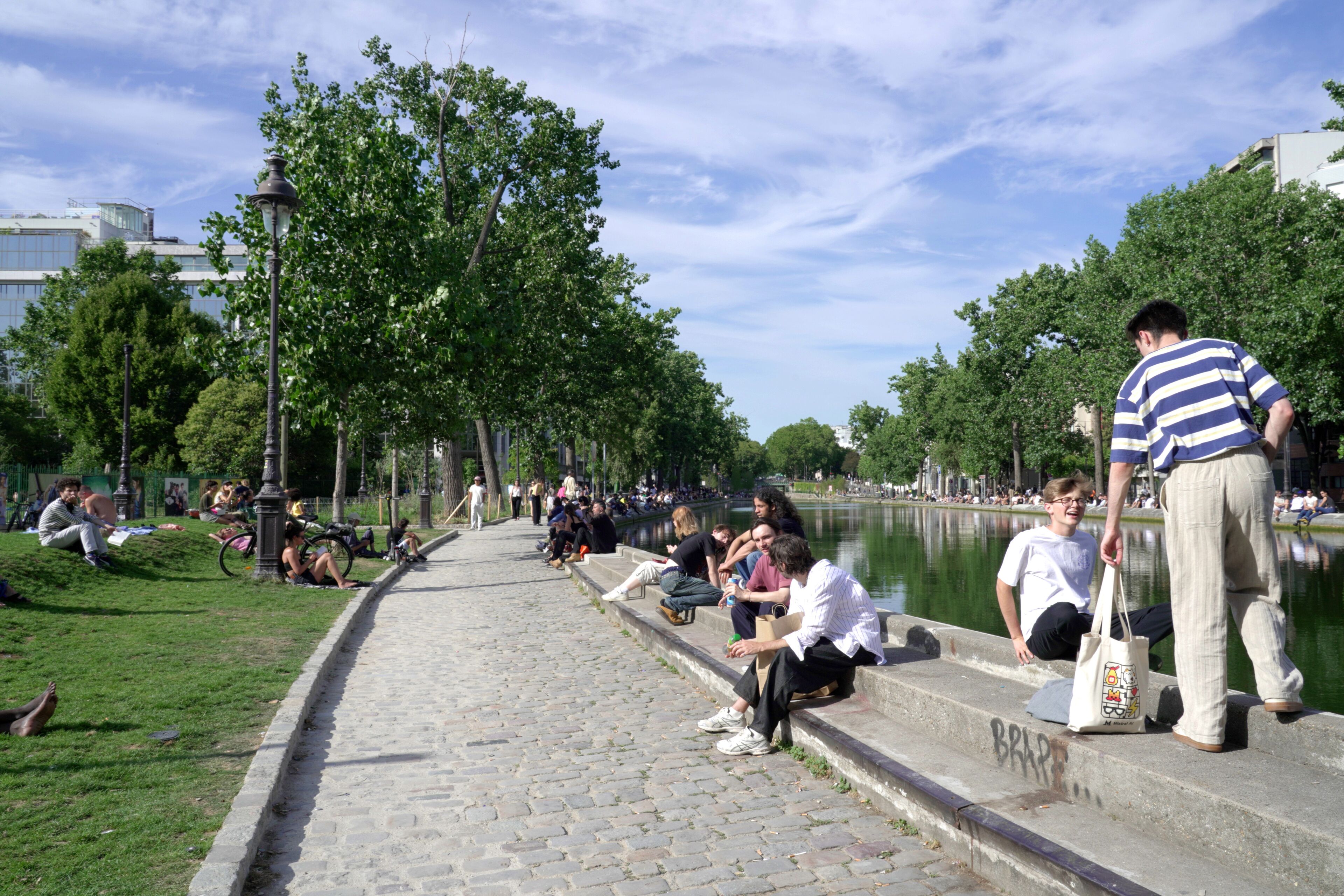 A park with people sitting on steps and grass, a body of water, and trees.