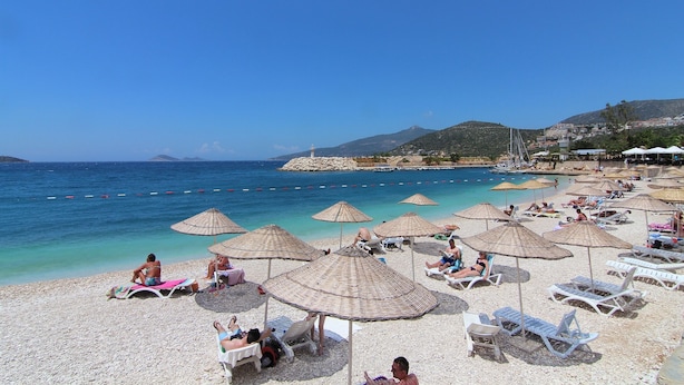 Une plage avec des chaises longues et des parasols, une eau d’un bleu limpide et un paysage montagneux en arrière-plan.