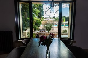 A dining table with a vase of flowers in a room with a large window overlooking a pool and garden. - RENTAL OF A MAS WITH POOL IN A VINEYARD IN FIELD CAMARGUE (Vauvert)