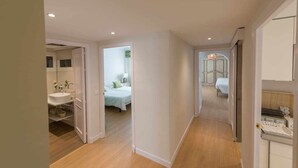 Een gang die leidt naar een slaapkamer met een bed, een nachtkastje en een bijzettafeltje. Links is een badkamer zichtbaar met een wastafel en spiegel.