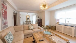 Een woonkamer met een beige bank, een houten salontafel en een eethoek met een kroonluchter.