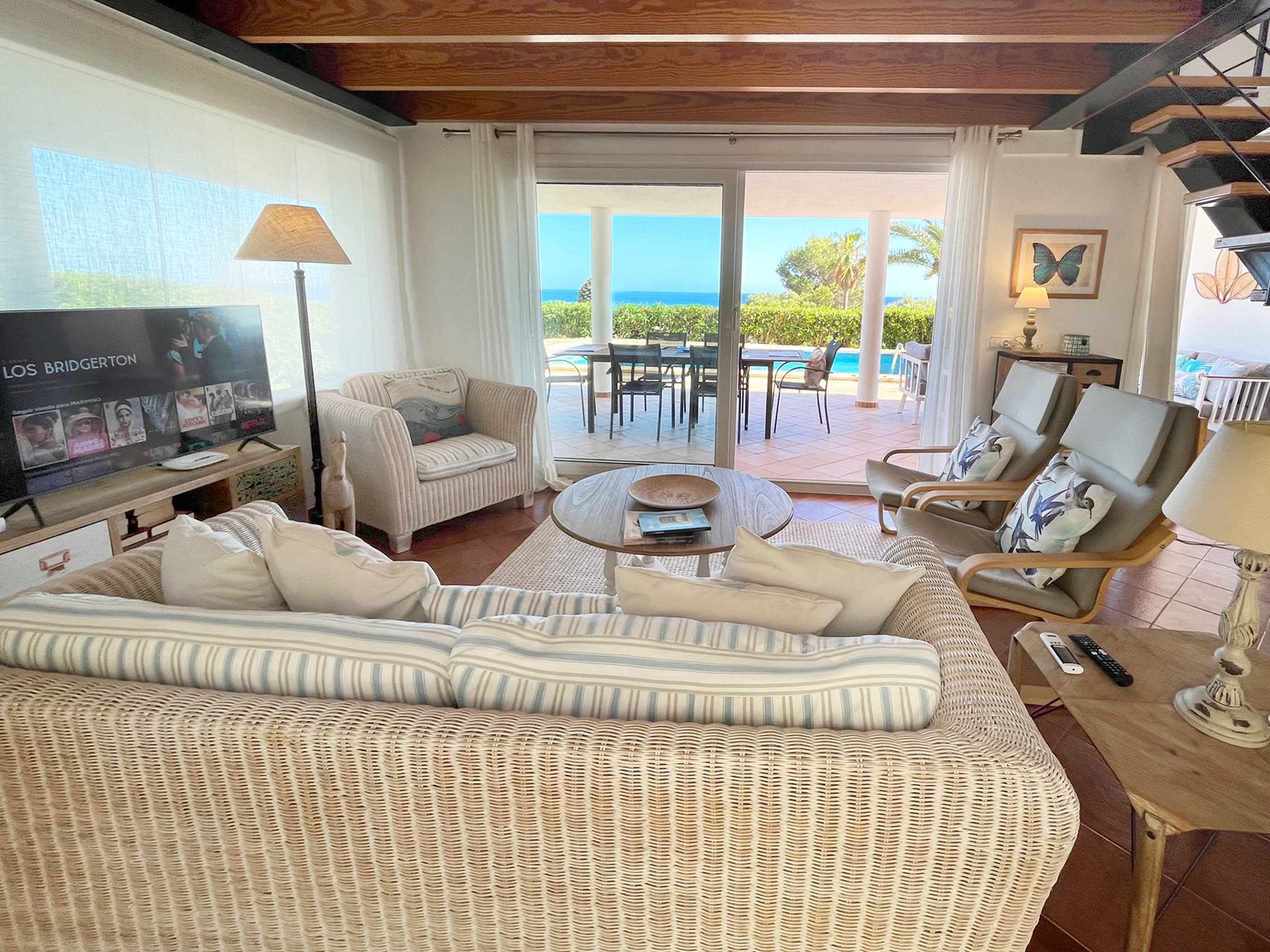 A living room with a large sofa, a coffee table, and a TV. There is a balcony with a pool and outdoor furniture.