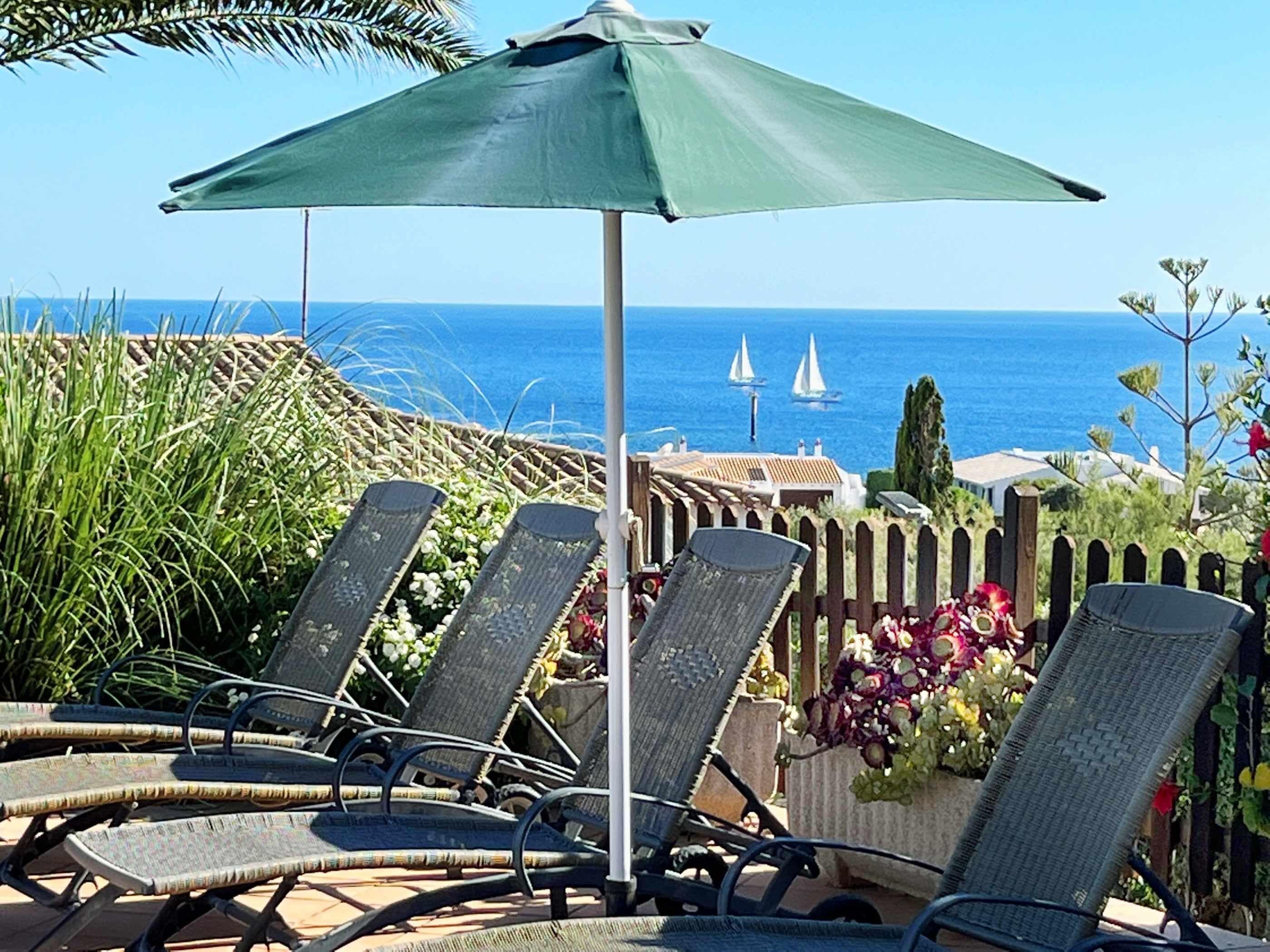 Een terras aan zee met ligbedden en een parasol, met uitzicht op de zee en boten.