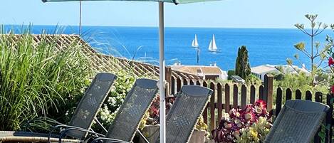 Een terras aan zee met ligbedden en een parasol, met uitzicht op de zee en boten.