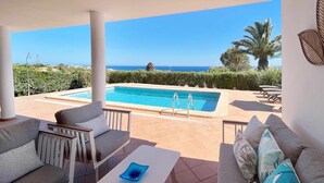 Area relax esterna coperta con piscina, mobili bianchi e vista sul mare.