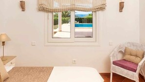 Una camera da letto con un letto, una sedia, una finestra vista piscina e vegetazione, e lampade a parete.
