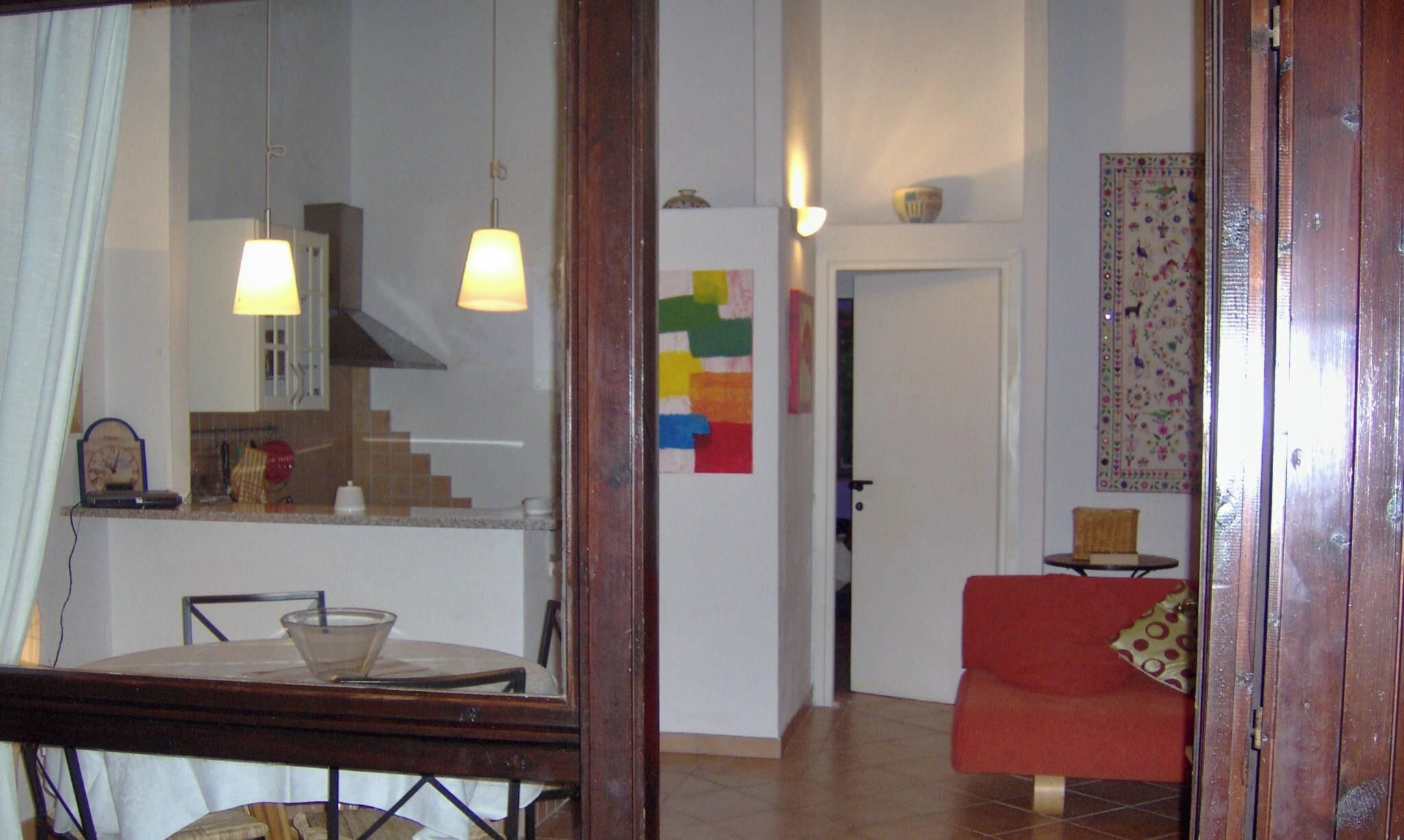 A dining area with a table and chairs, a kitchen in the background, a red sofa, and a door leading to another room.