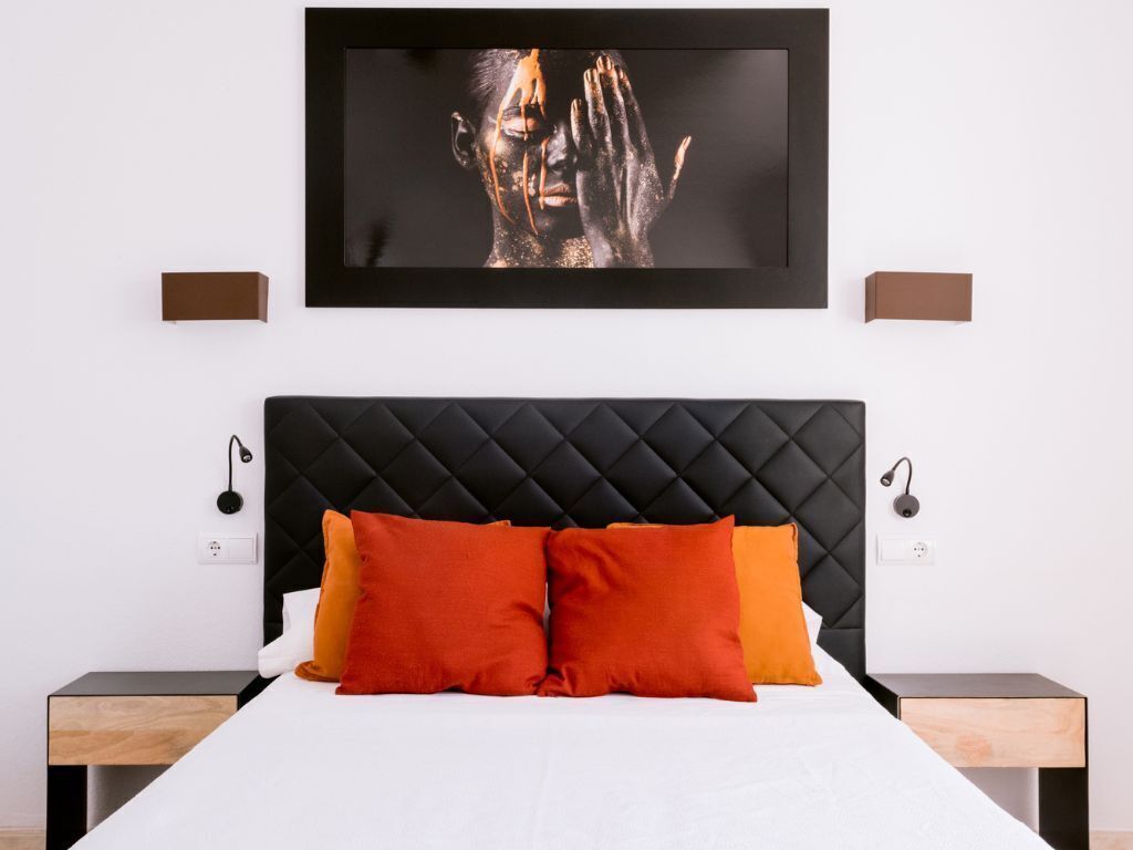 Un dormitorio moderno con un cabecero tapizado negro, una colcha blanca y dos almohadas naranjas.