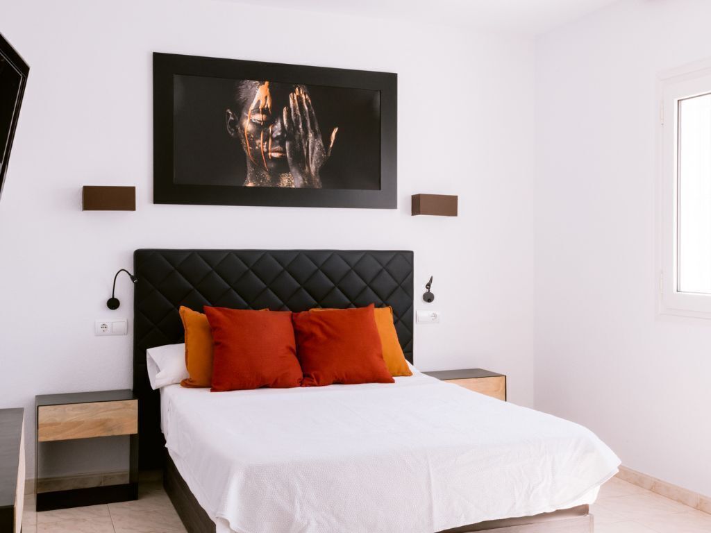 Un dormitorio con cabecera negra, ropa de cama blanca y dos almohadas naranjas.