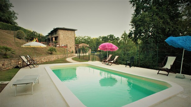 Piscina com espreguiçadeiras e guarda-sóis, cercada por um muro de pedra e vegetação.
