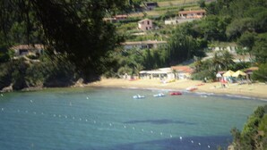 A coastal area with clear water, a sandy beach, and several small boats. There are buildings in the background, some with red roofs. - Colle Paradiso Island of Elba from the tranquility of nature to the sea in 10 minutes (Capoliveri)