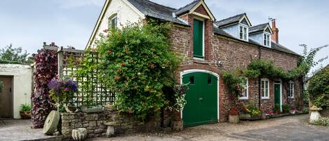 Une maison traditionnelle en pierre, avec une porte verte, un mur de pierre et une clôture en bois ornée de plantes grimpantes.