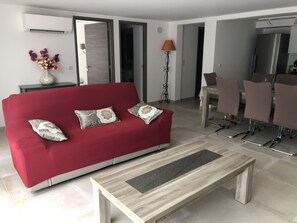 A modern living room with a red sofa, two decorative pillows, a coffee table, a dining area with chairs, and a kitchen visible through an open doorway.