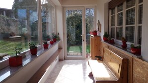Un couloir baigné de lumière, avec un banc en bois, des plantes en pot sur le rebord de la fenêtre et une vue sur un jardin par une porte vitrée.