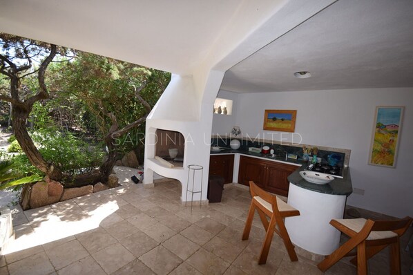A kitchen with a built-in island, a dining area with two wooden chairs, and a view of a garden with trees and rocks. - Villa Sassifraga: just few steps from the beach (Portobello Di Gallura)