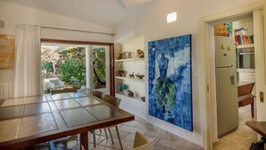 Un coin repas avec une table et des chaises, une cuisine équipée d’un réfrigérateur et comprenant des livres, et un grand tableau abstrait accroché au mur.