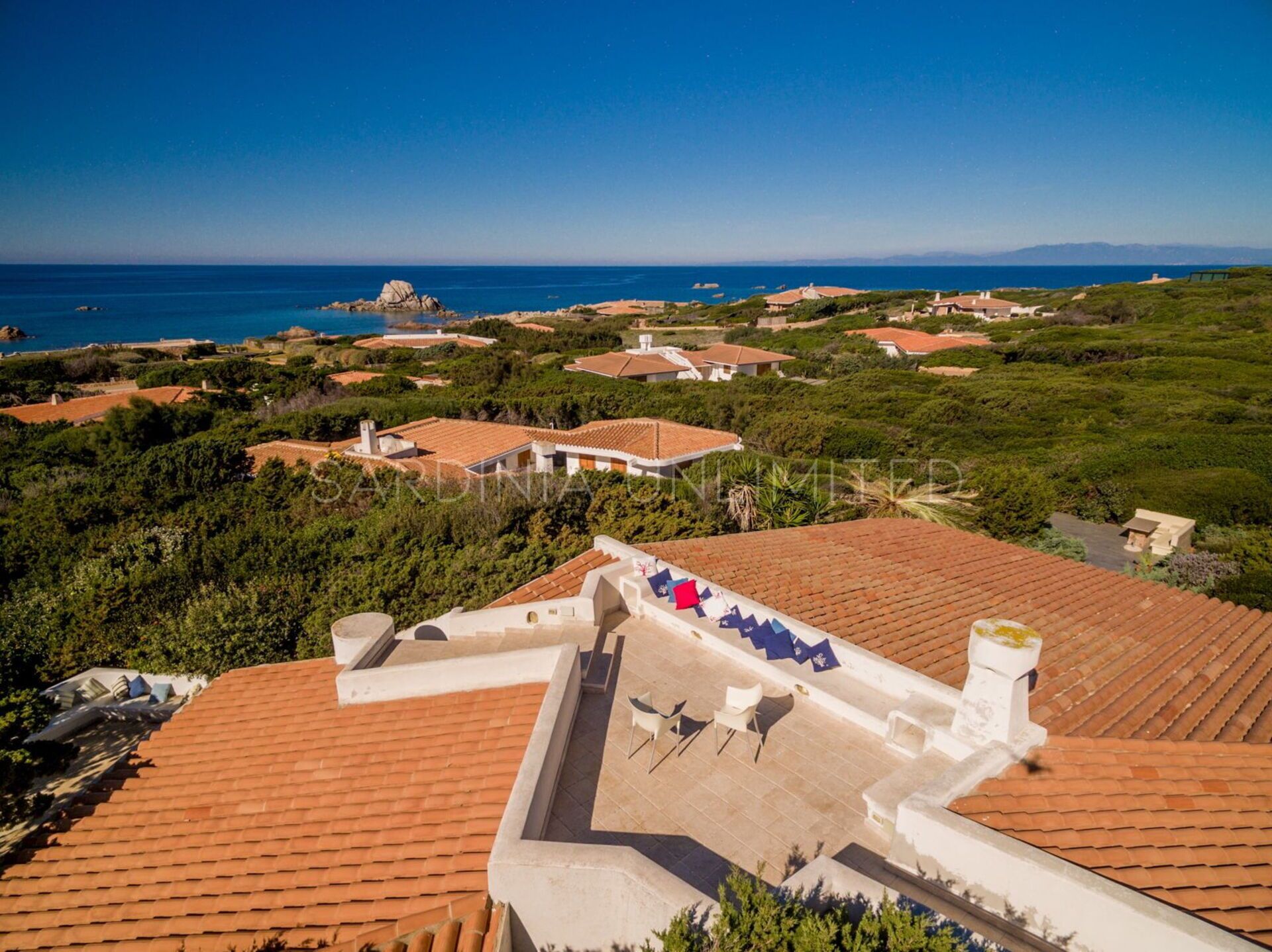 A coastal residential area with terracotta-tiled roofs, a patio with outdoor furniture, and a view of the sea and distant mountains.