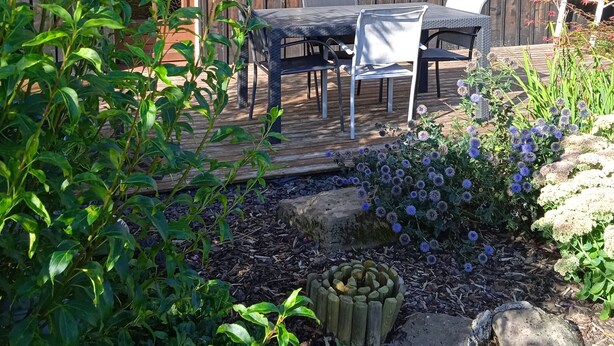Ein Holzgebäude mit einer Terrassenanlage, komplett mit Tisch und Stühlen, umgeben von Pflanzen und Blumen.