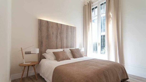 A modern bedroom with a large bed, a bedside table with a lamp, and a view of the outside through a window. - Stylish Apartment C in city center (Barcelona)
