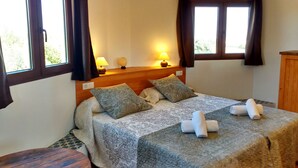 Una cama bien hecha con ropa de cama y almohadas estampadas, una mesita de noche con lámparas, y vistas a través de la ventana.