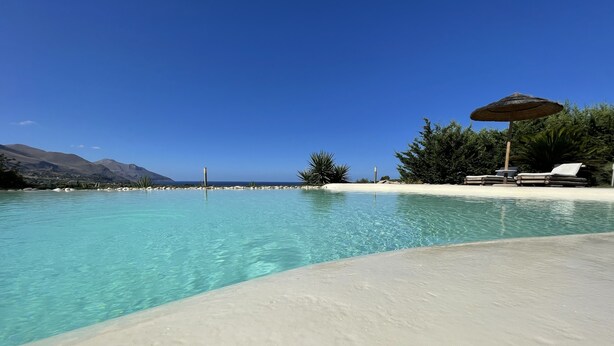 A clear pool with a sandy beach, lounge chairs, and an umbrella.