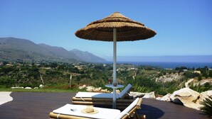 A wooden deck with a thatched umbrella, lounge chairs, and a view of the sea and mountains.