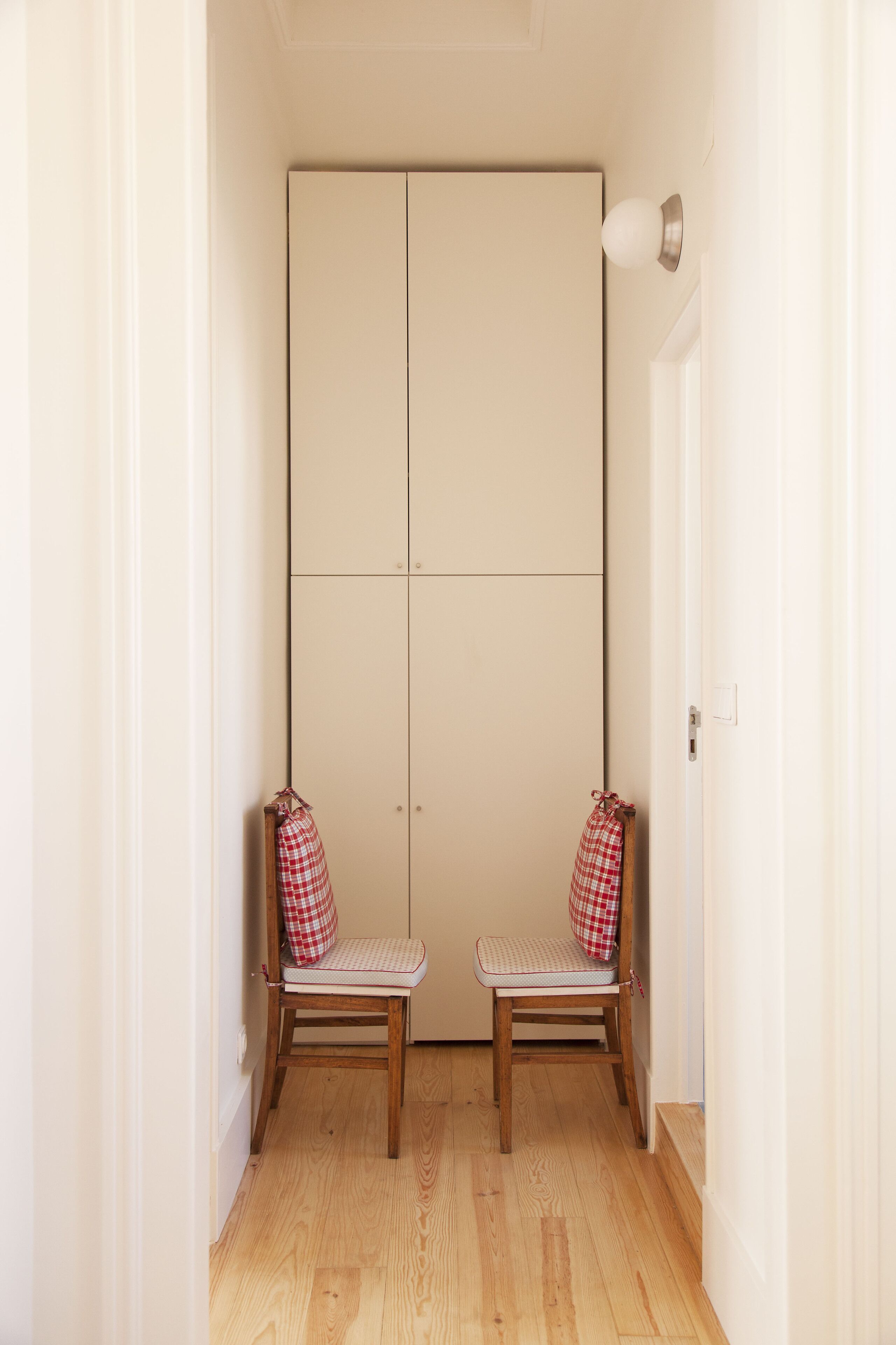 A hallway with two wooden chairs with checkered cushions, a beige wardrobe, and a light switch on the wall.