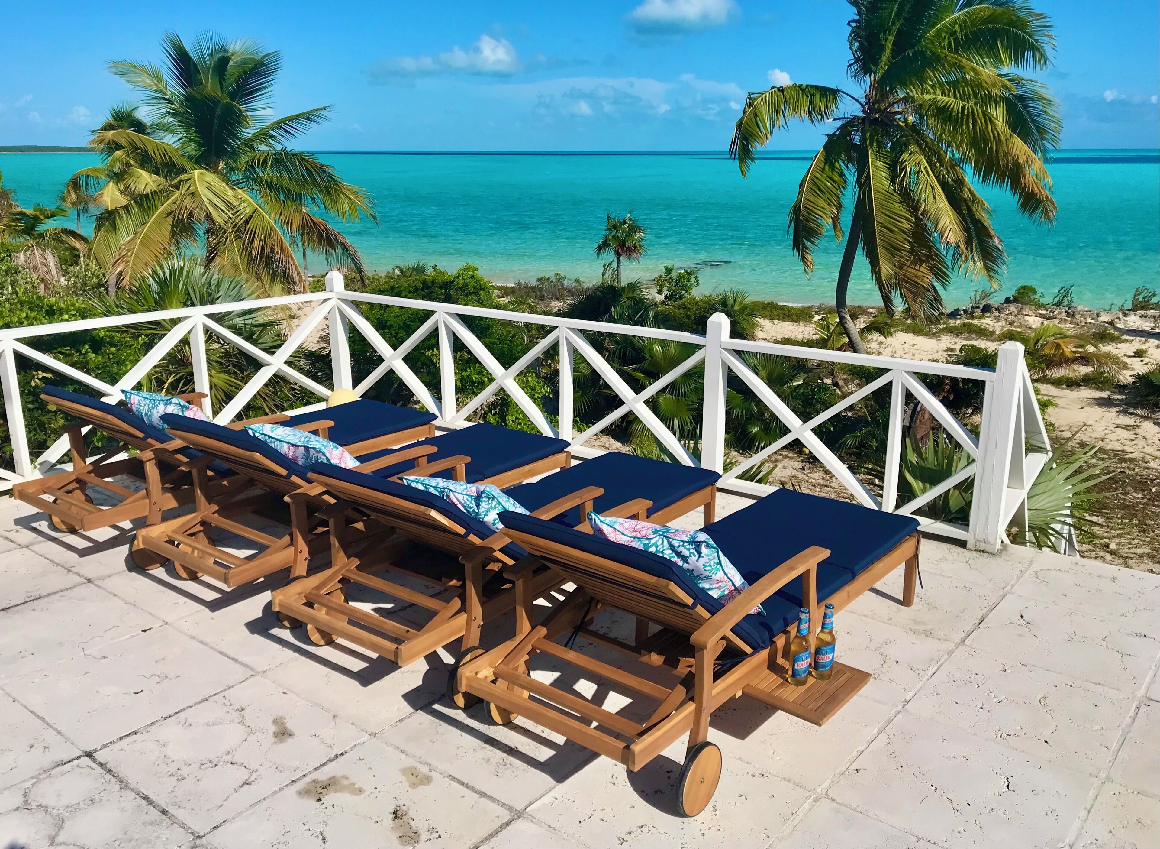 Un espace de détente en bois avec des coussins bleus, une balustrade blanche et une vue sur la mer turquoise et les palmiers.