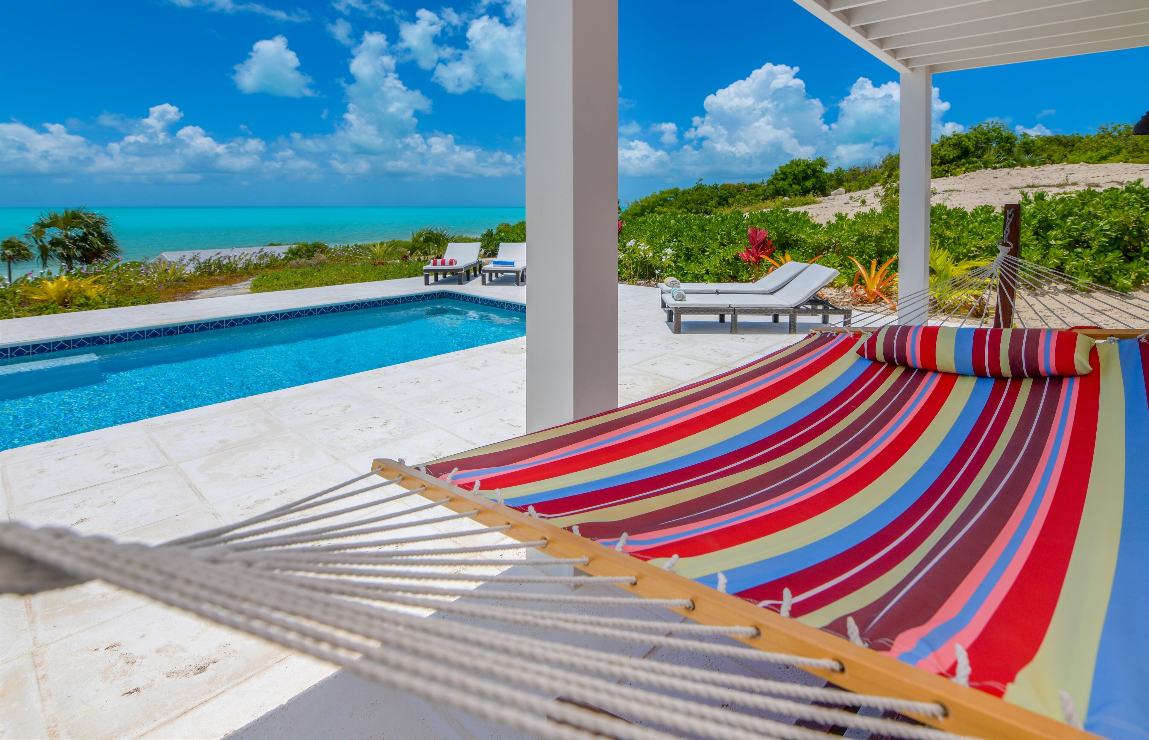 A hammock with a colorful striped fabric, a pool, and a view of the ocean and greenery.