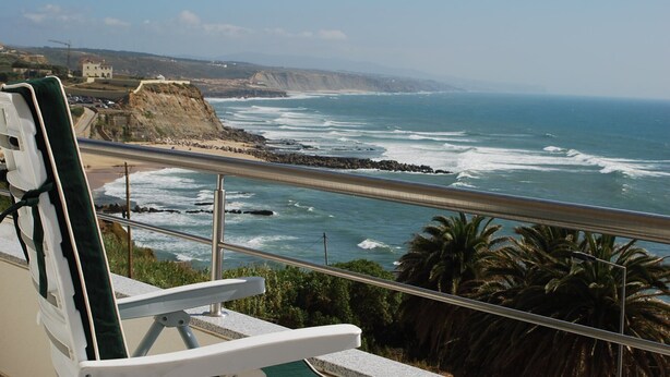 Vista para o mar a partir de uma varanda com guarda-corpo, com vista para a costa rochosa e o oceano.