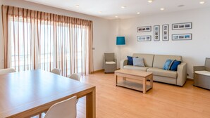 Uma sala de estar moderna com piso de madeira, um sofá bege, uma mesa de centro e uma mesa de jantar com cadeiras. Há luminárias no teto e quadros na parede.