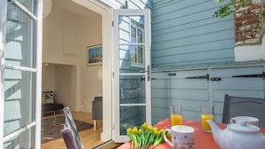 An open sliding glass door leading to a dining area with a table set for breakfast, including a teapot, two glasses of orange juice, and a bouquet of tulips.