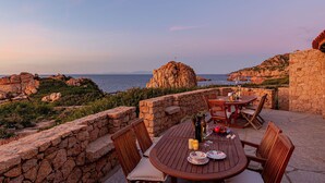 An outdoor dining area with stone walls, a table set for two, and a view of the sea and rocky landscape. - Villa Sfumature del Melograno: private pool in front of the sea (Costa Paradiso)