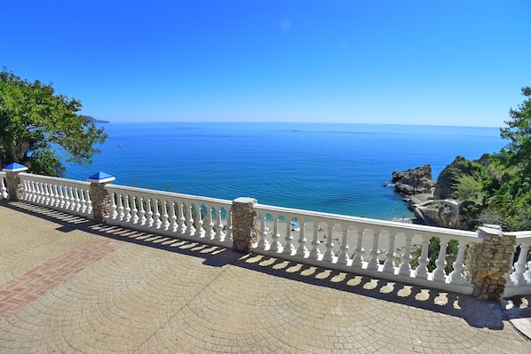 Una terraza frente al mar con barandilla blanca, pilares de piedra y un paseo empedrado con vistas al océano.