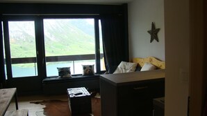 Ein Zimmer mit großem Fenster, das auf einen See und Berge blickt, eine Holzbank mit Kissen, ein Kuhfellteppich und ein Bett mit einem Holzkopfstück.