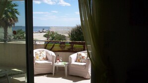 Un balcon avec deux fauteuils blancs dotés de coussins, une petite table et une bouteille de vin. On peut admirer la vue sur la plage à travers la porte vitrée.