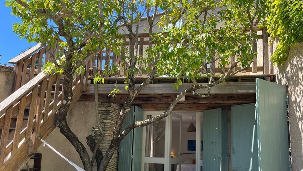A hammock tied between two trees in a courtyard with a wooden staircase and a building with open doors.