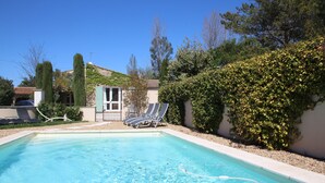 A swimming pool with clear blue water, surrounded by a hedge and a building with a tiled roof.