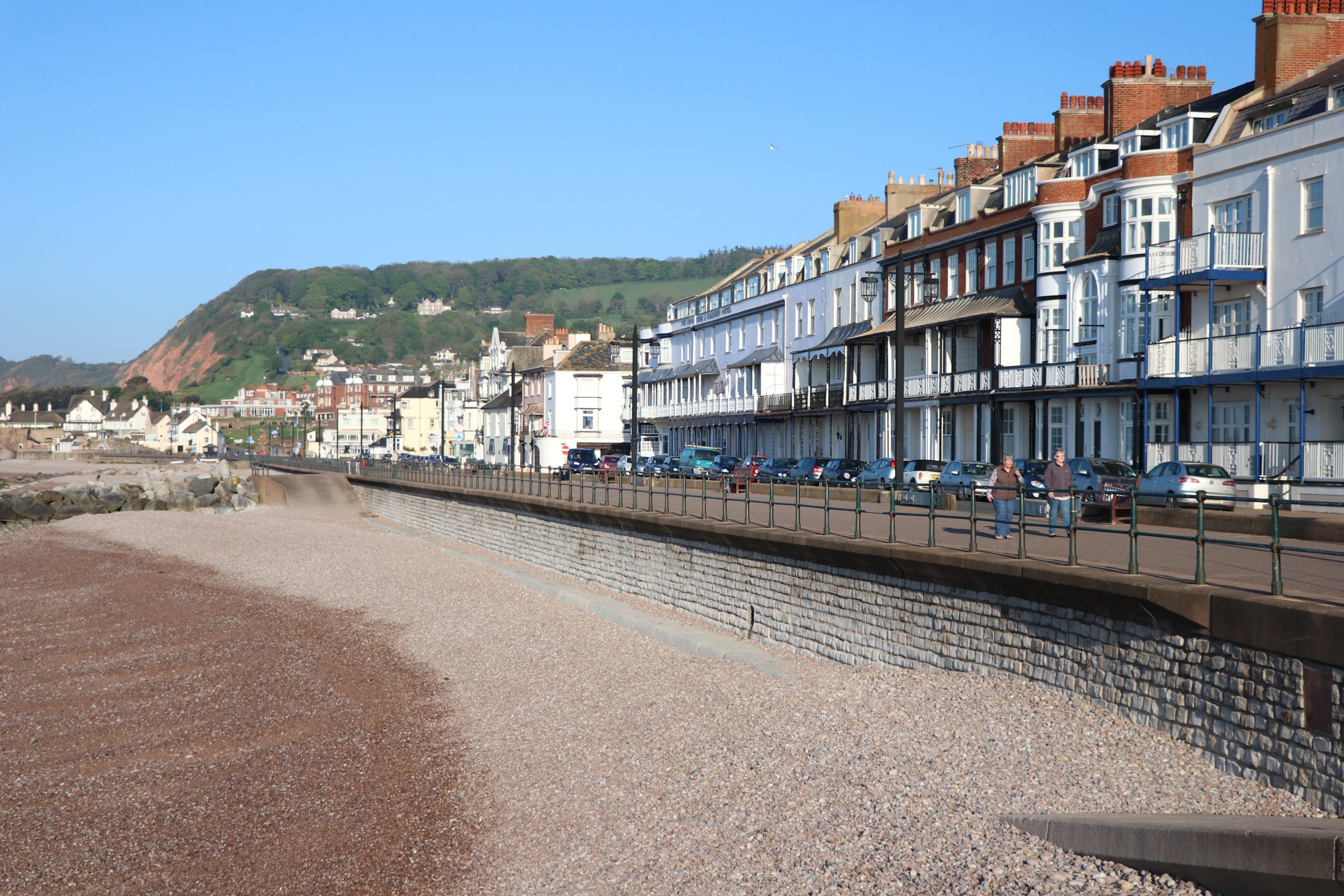 A coastal promenade with terraced houses, a pebble beach, and a clear sky.